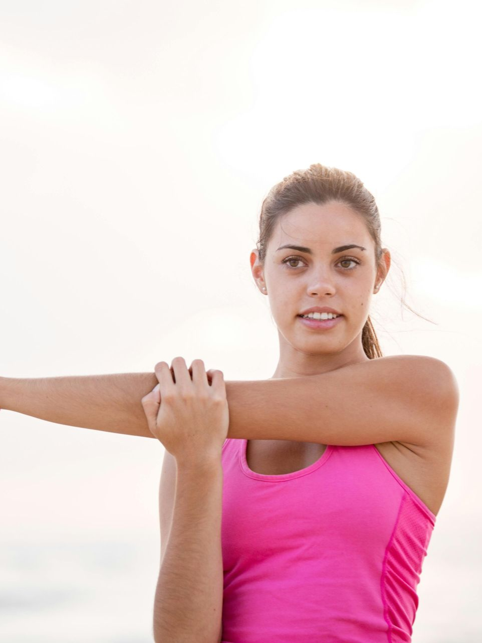 A person in a bright pink tank top performs a cross-body shoulder stretch outdoors against a bright, hazy background.