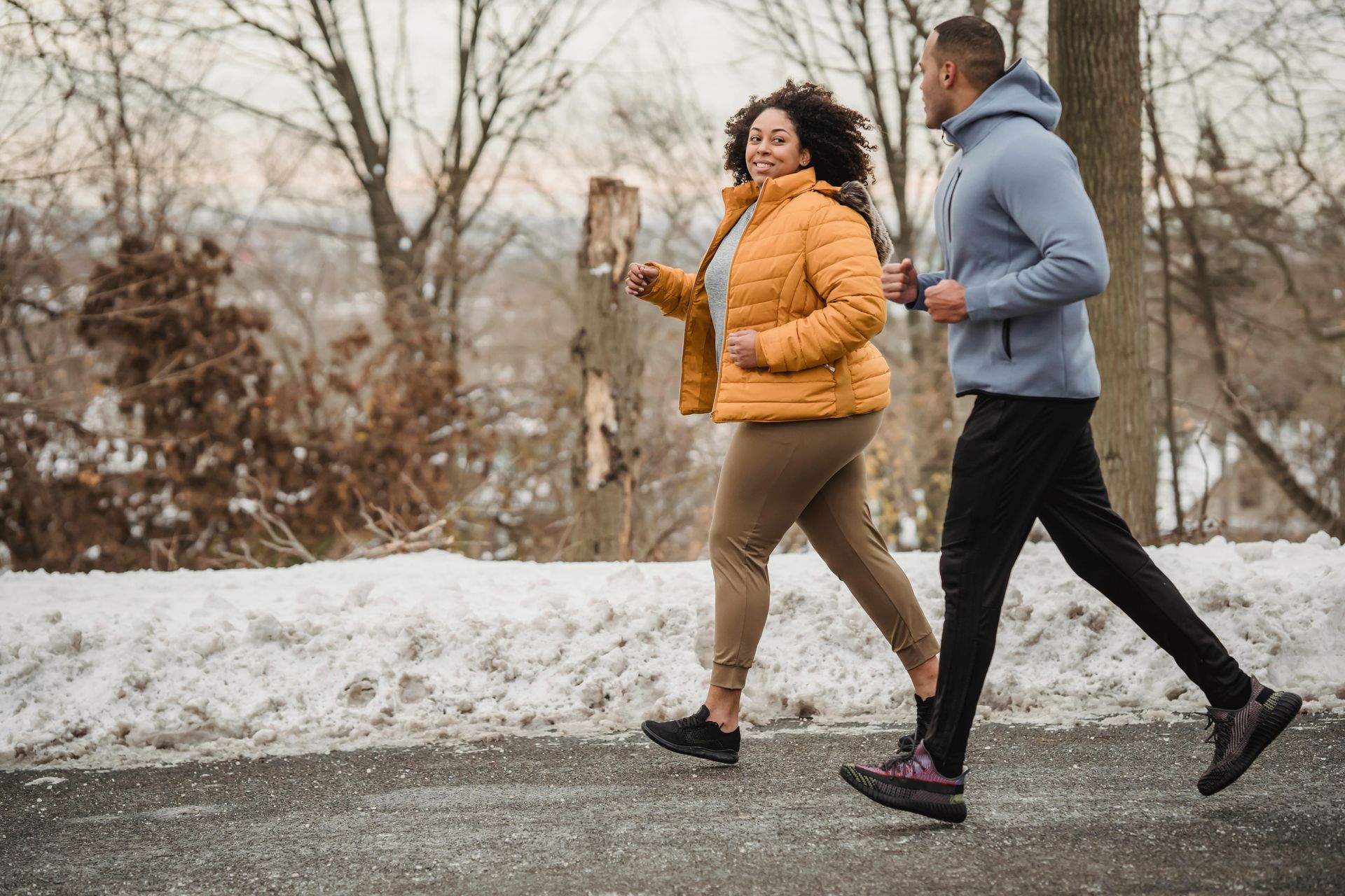 Two people jogging on a snow-lined path. One wears a yellow jacket, the other a blue hoodie.