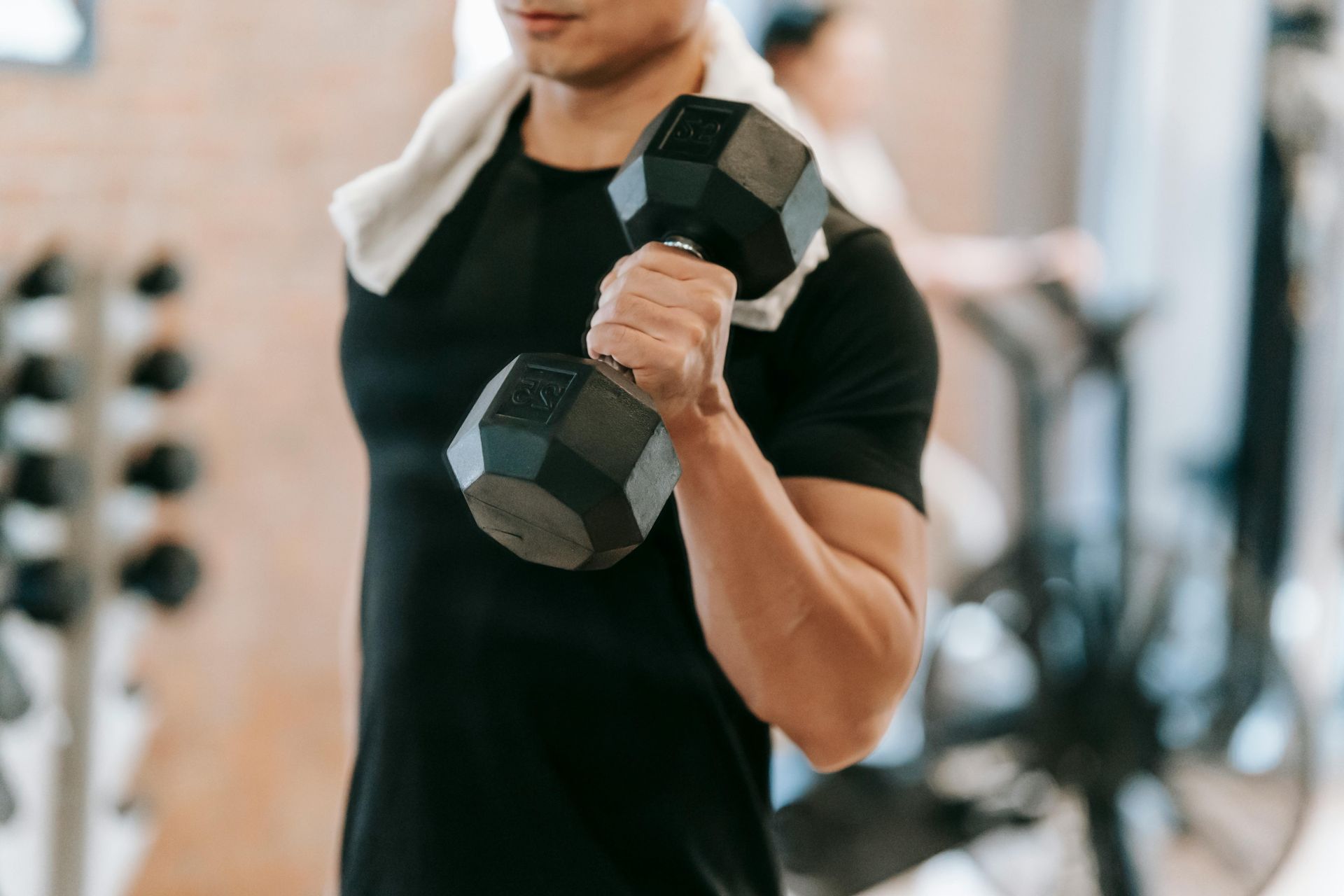 Man lifting a dumbbell in a gym; white towel around neck; black shirt.