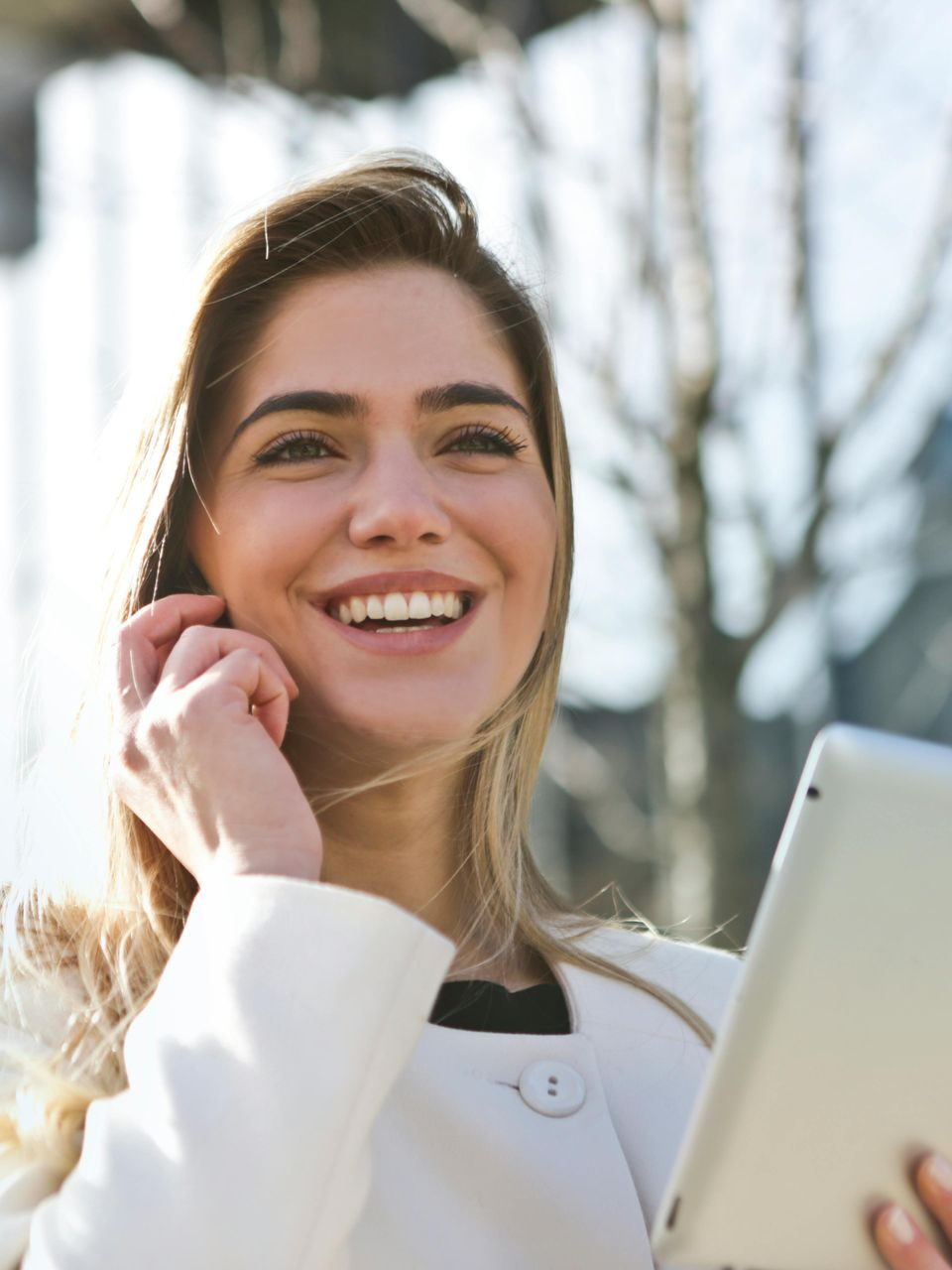 Smiling person in a white jacket talking on a phone and holding a tablet outdoors.