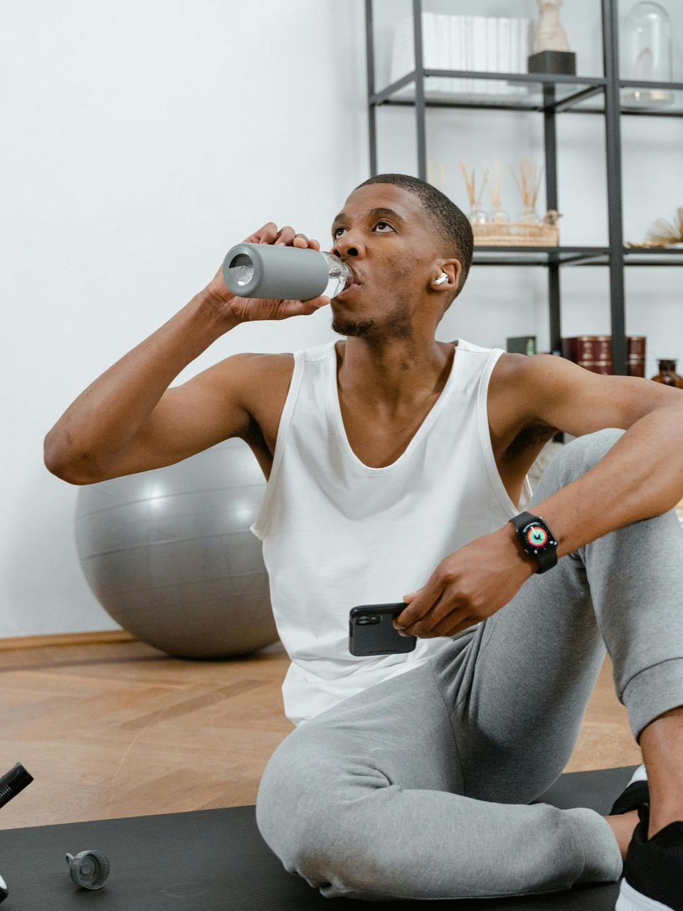 An individual sits on a gym mat drinking from a grey bottle while holding a smartphone in a home fitness space.