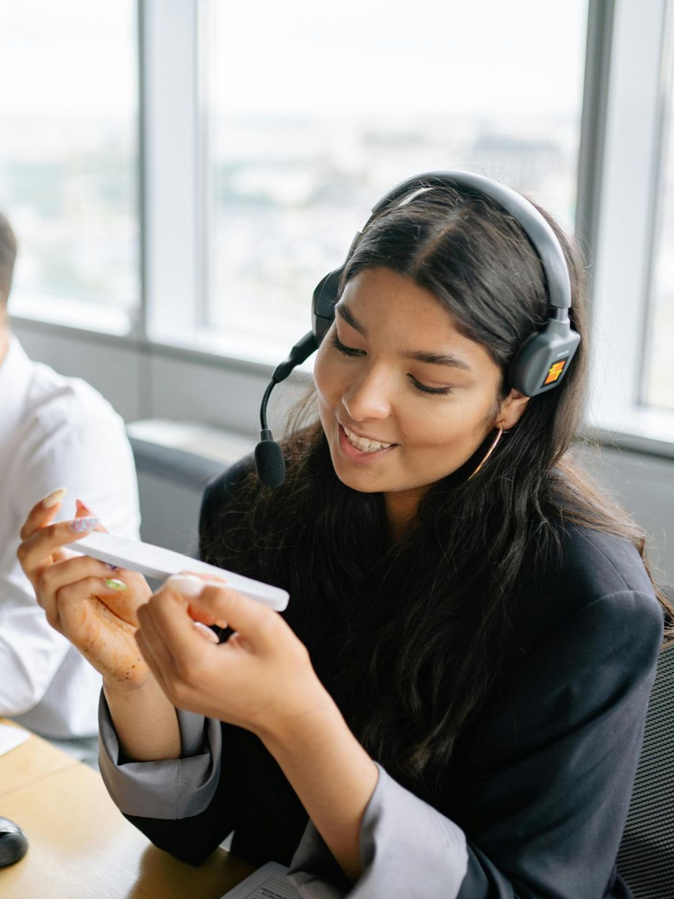 A person wearing a headset holds a small, white rectangular device while smiling in an office setting.