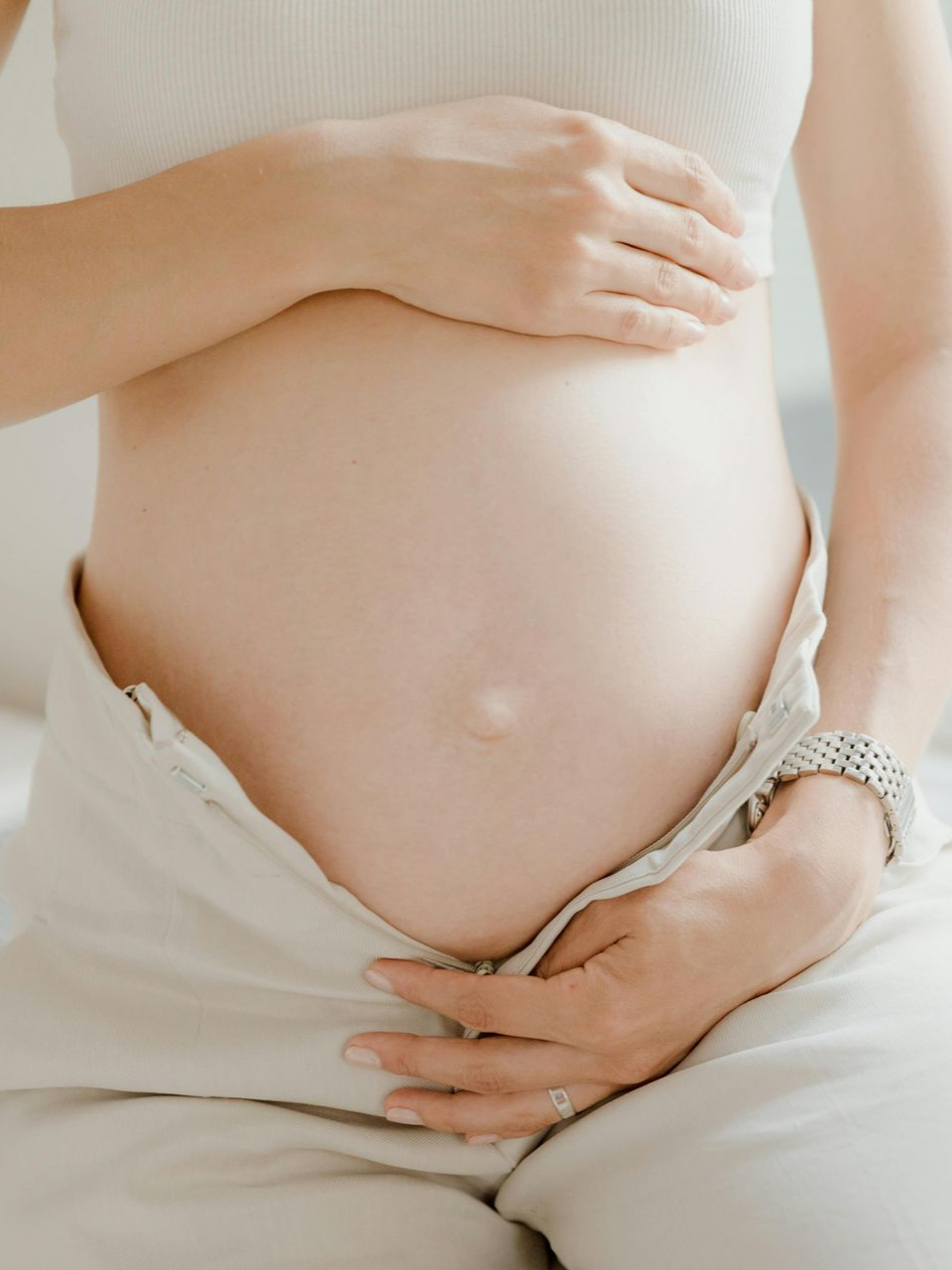 A pregnant person wearing light-colored clothing holds their stomach while sitting near a window.