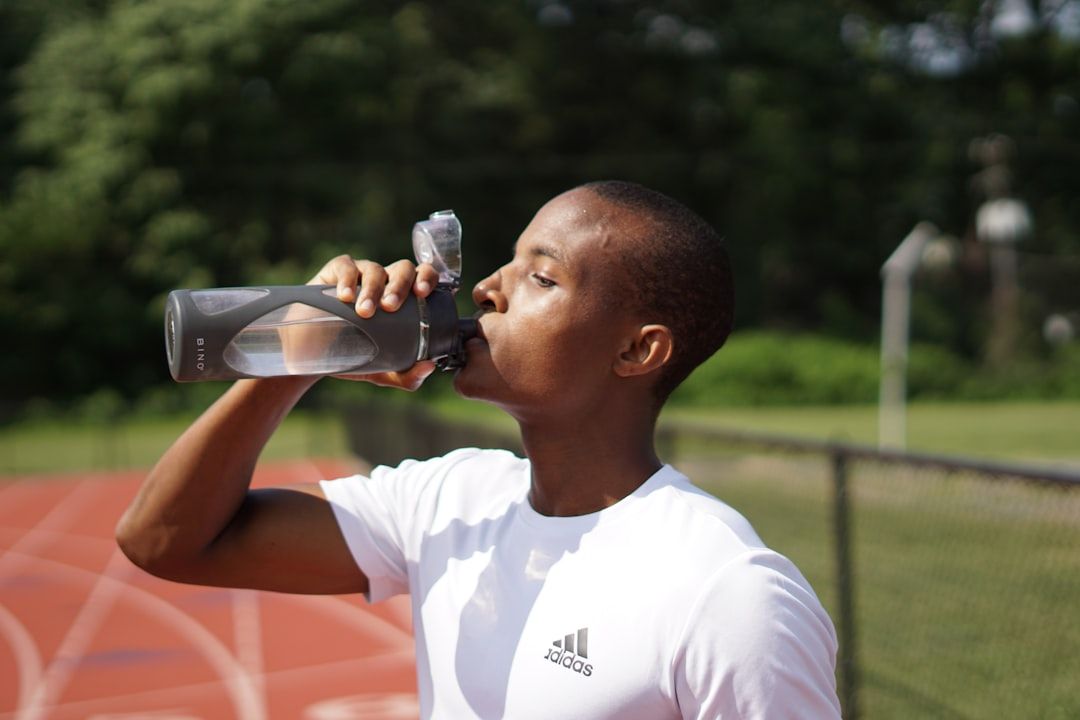 A person in a white Adidas t-shirt drinks from a water bottle while standing on an outdoor track.