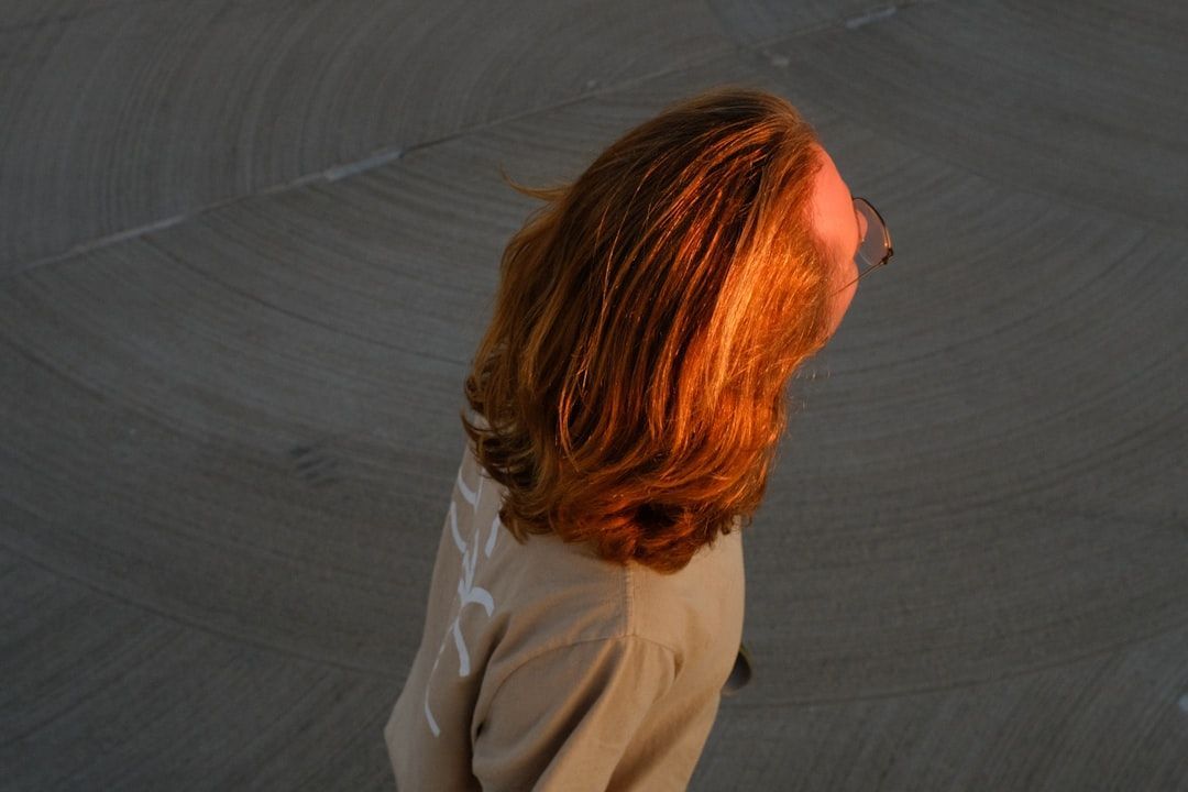 A person with wavy, reddish-brown hair illuminated by warm sunlight, wearing a beige top against a dark, curved background.
