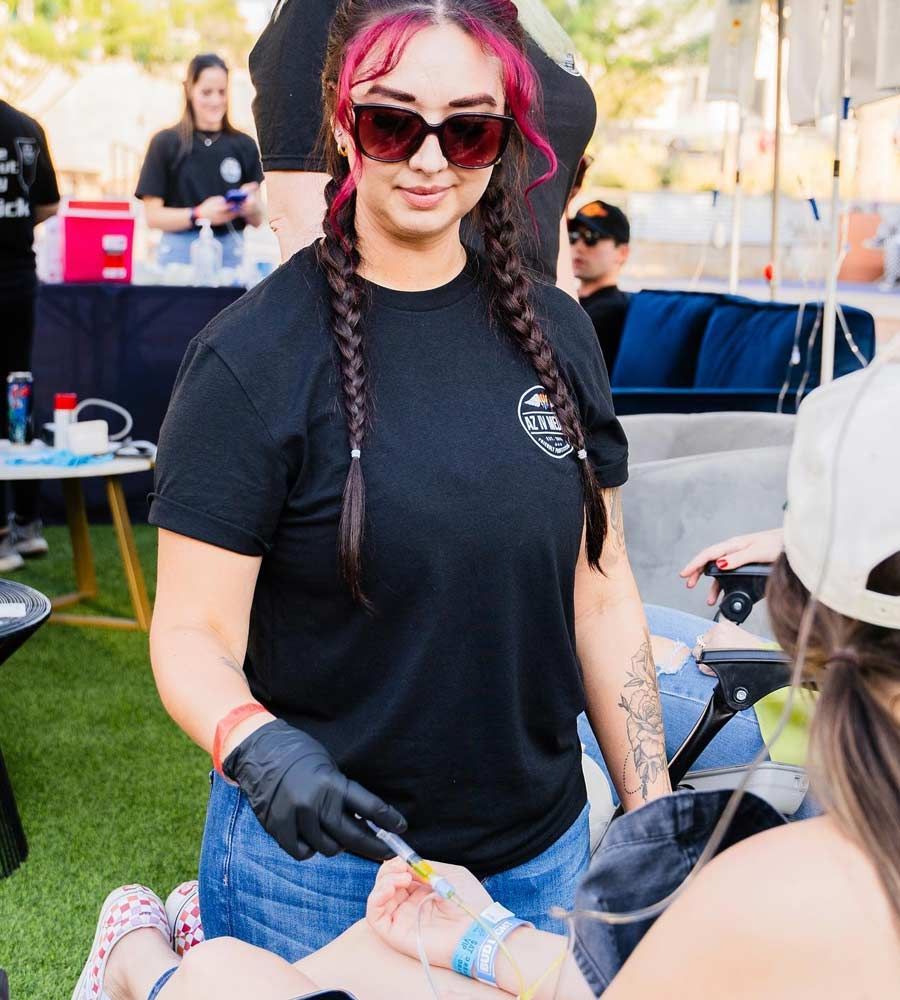 A person wearing black gloves and sunglasses administers an IV drip to someone seated at an outdoor event.