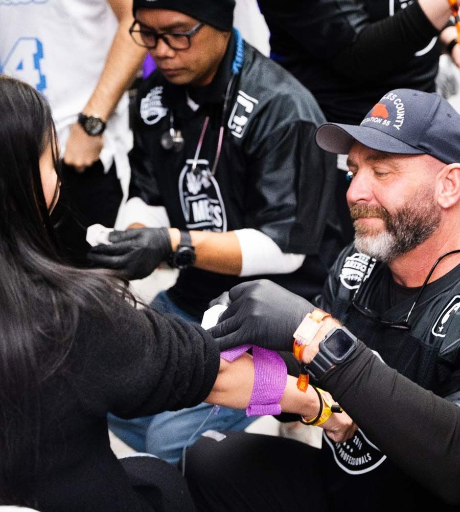 A medical professional applies a purple tourniquet to a person's arm while an assistant prepares supplies nearby.