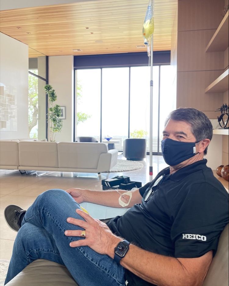 A person wearing a black mask and shirt sits on a sofa, receiving an IV infusion in a bright, modern living room.