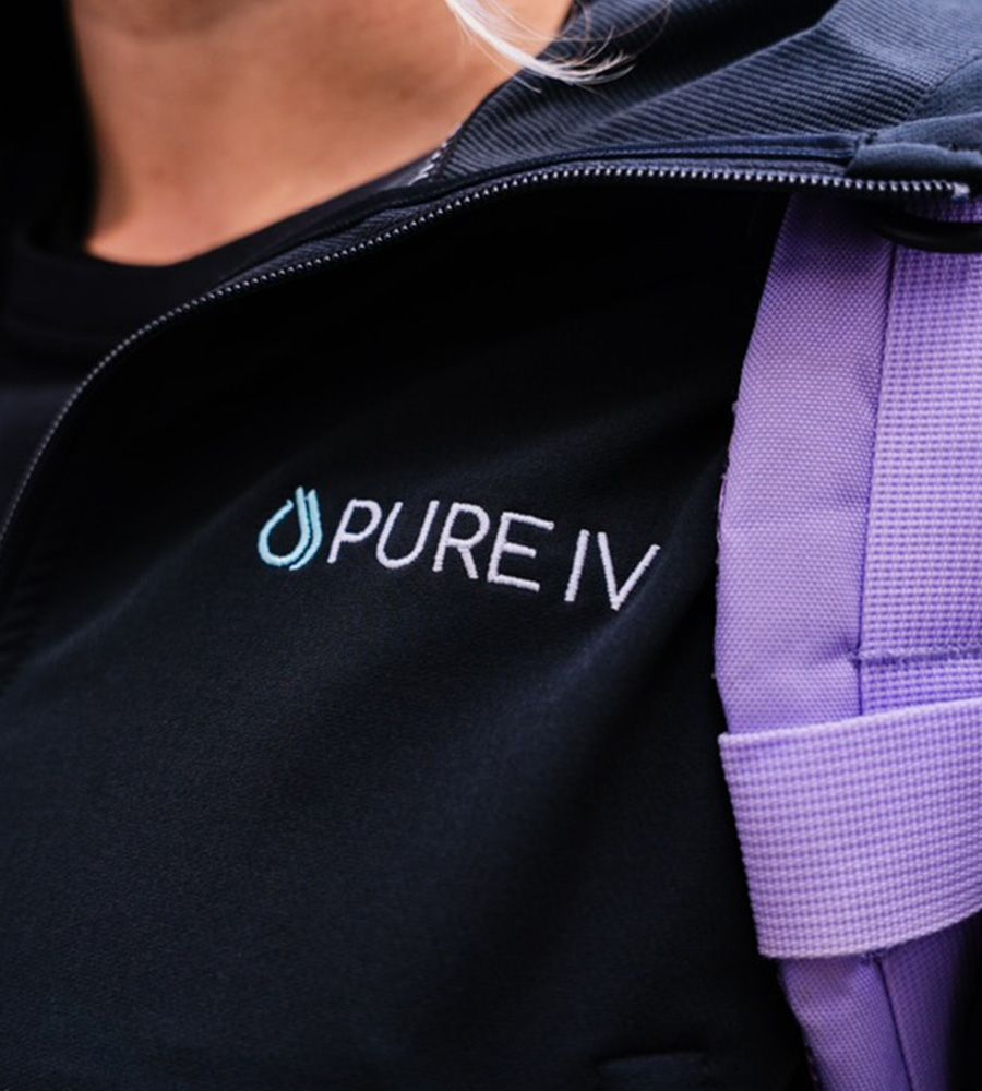 Close-up of the PURE IV logo embroidered in white and light blue on a professional black staff uniform jacket, highlighting quality branding and staff attire.