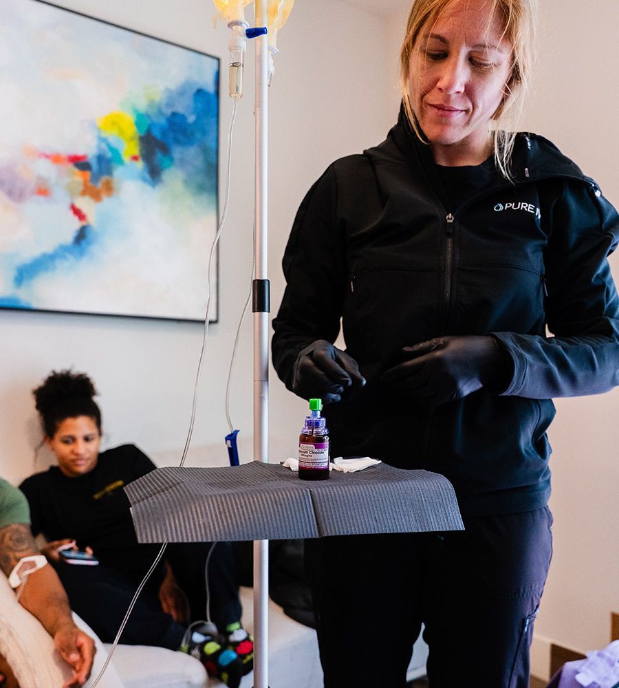 Professional PURE IV nurse in a black uniform and black gloves carefully preparing a customized vitamin vial on a sterile tray attached to the IV pole, with clients relaxing during their mobile infusion in the background.