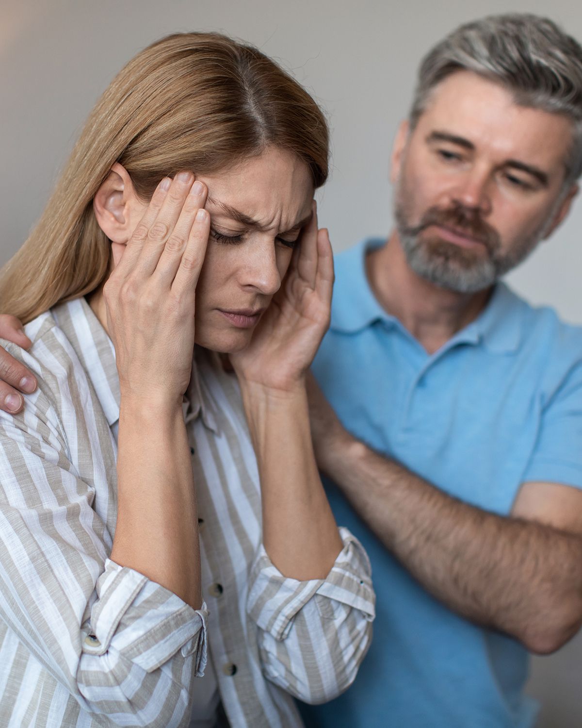 A person with eyes closed holds their head in pain while another person places hands on their shoulders in support.