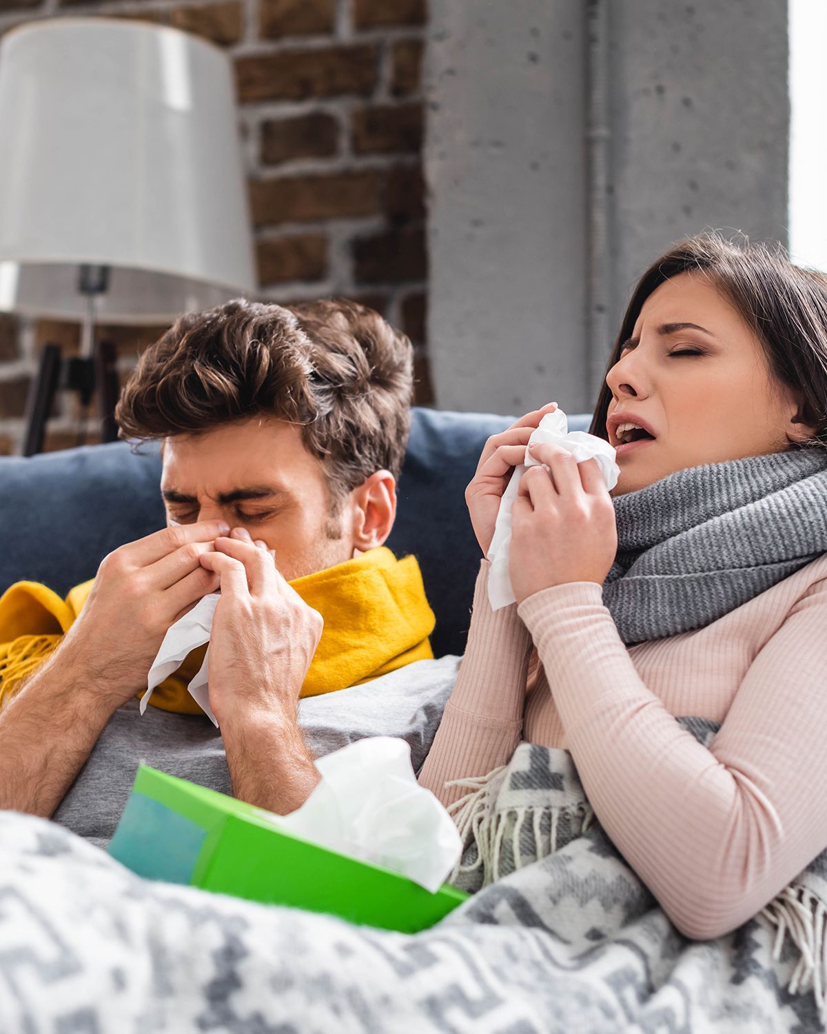 Two people with colds, wrapped in blankets and scarves, use tissues while sitting on a couch.