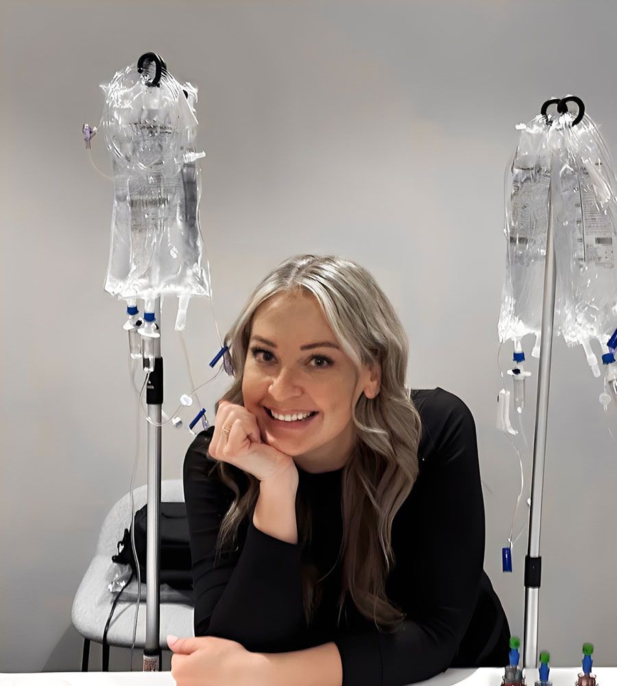 Smiling blonde woman posing confidently behind a table featuring two IV therapy poles and hanging hydration bags.