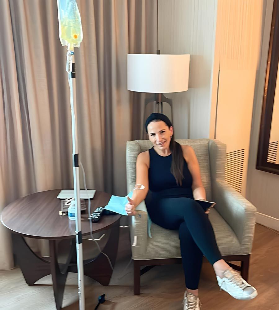 Smiling woman receiving an IV hydration drip while relaxing in a chair in a hotel room during travel.