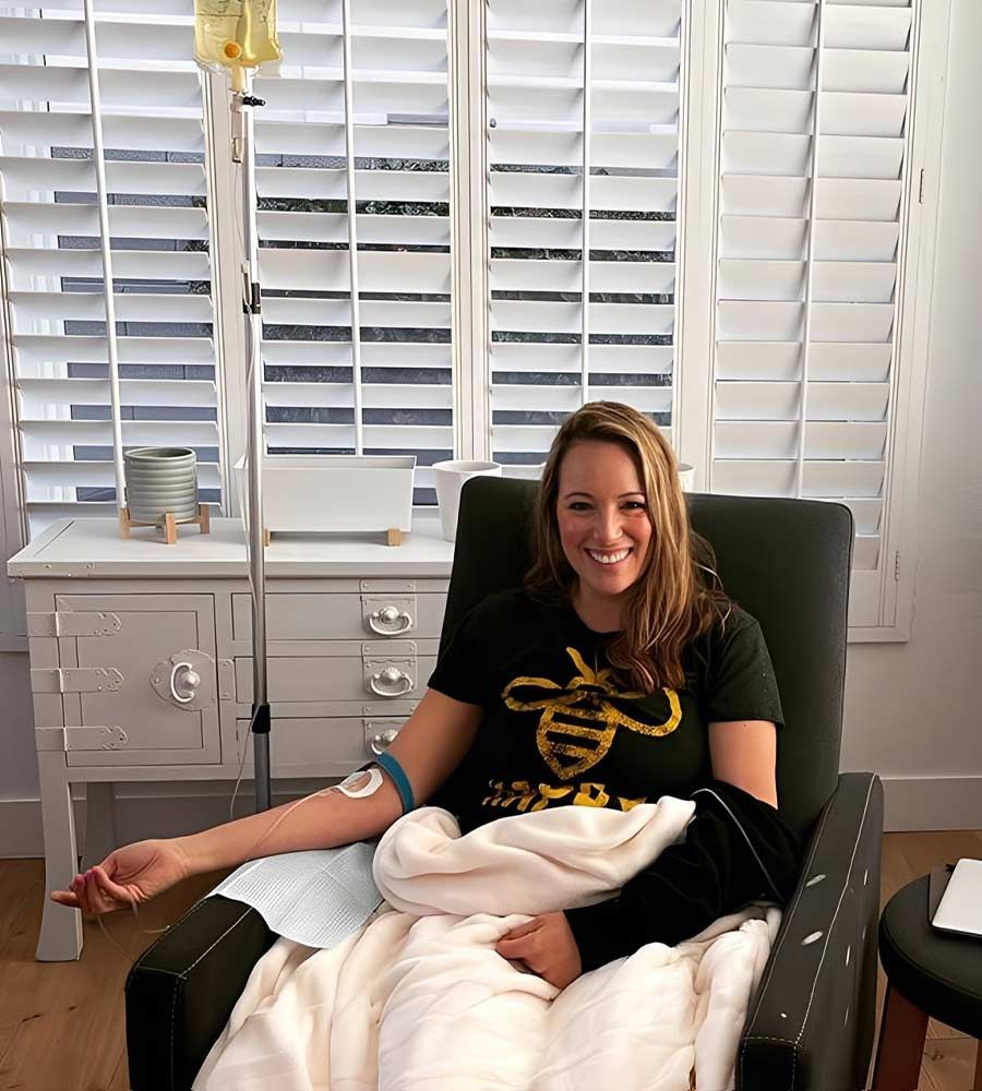 Smiling woman wearing a bee shirt relaxing in a chair and receiving an in-home IV vitamin drip.