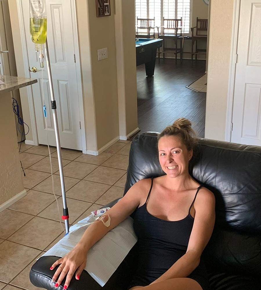 Smiling woman relaxing in a black leather chair while receiving an IV hydration treatment at home with a pool table visible.