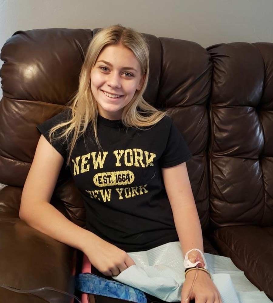 Smiling young blonde woman sitting on a brown leather couch receiving an IV treatment in her hand.