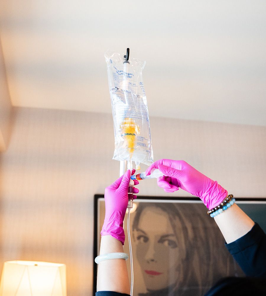 Close-up of a professional in bright pink gloves injecting customized nutrients from a syringe into a hanging IV fluid bag, emphasizing sterile and precise preparation for the infusion.