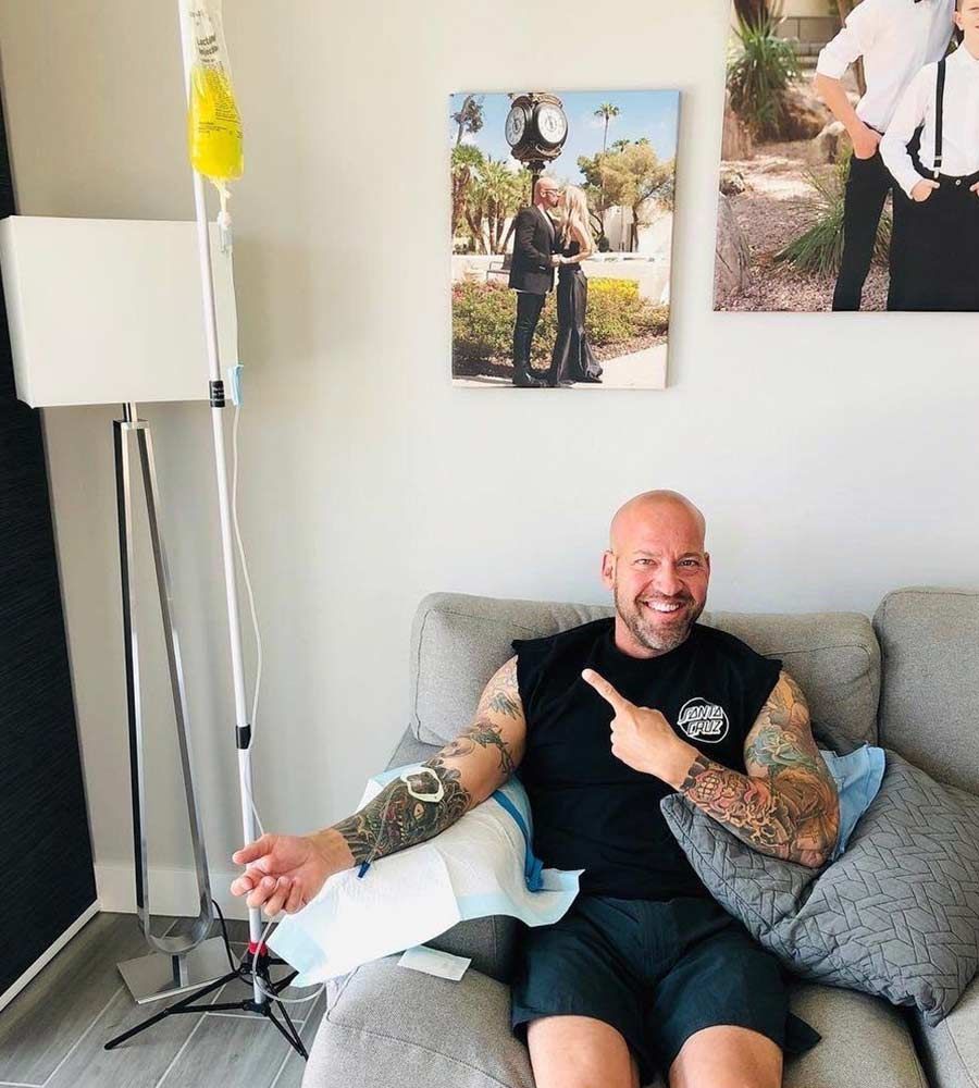 Smiling, tattooed man pointing to his arm while receiving an in-home IV vitamin infusion on a couch.