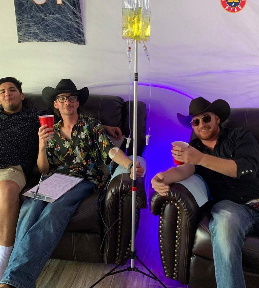 Three men in cowboy hats relaxing with red cups, one receiving a recovery IV hydration drip on a couch with purple lighting.