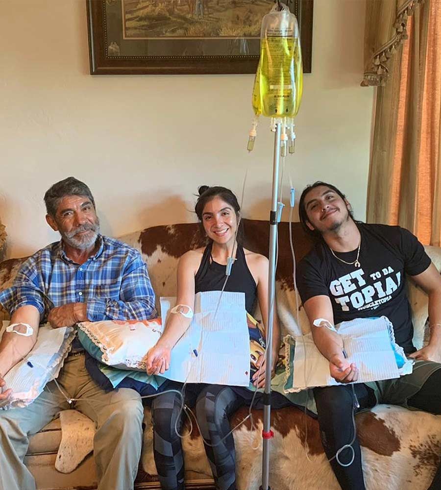 Three smiling people, appearing to be a family, sitting on a couch receiving simultaneous IV hydration treatments.