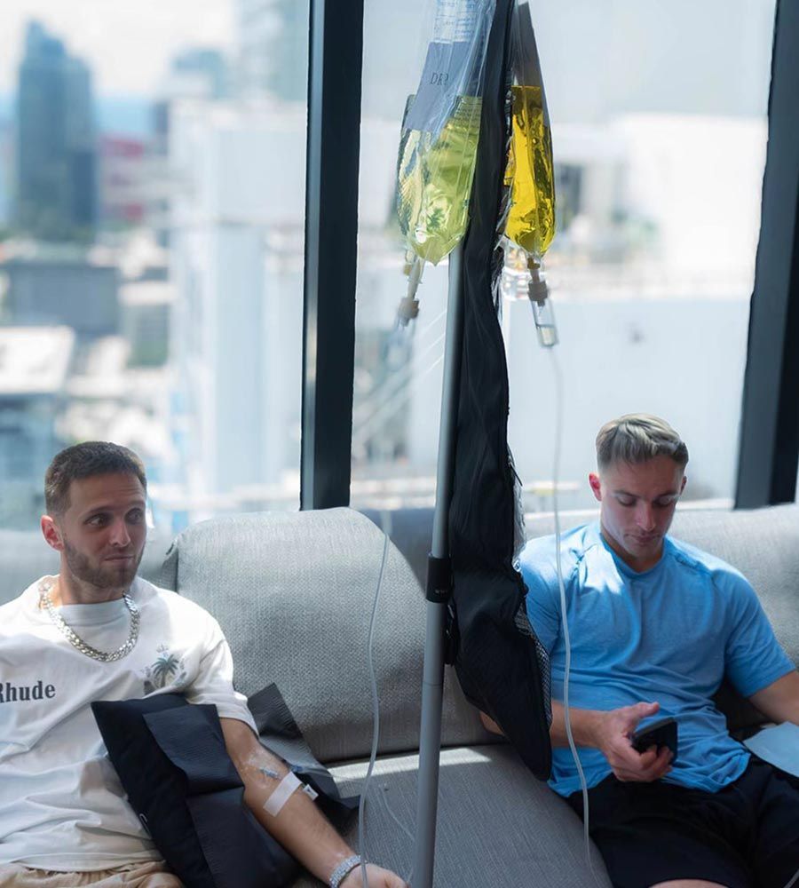 Two men receiving IV hydration drips while sitting on a couch in a high-rise with a city skyline view.