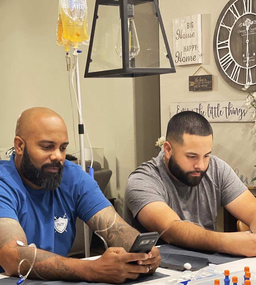 Two men sitting at a table checking their phones while receiving simultaneous IV vitamin therapy infusions at home.