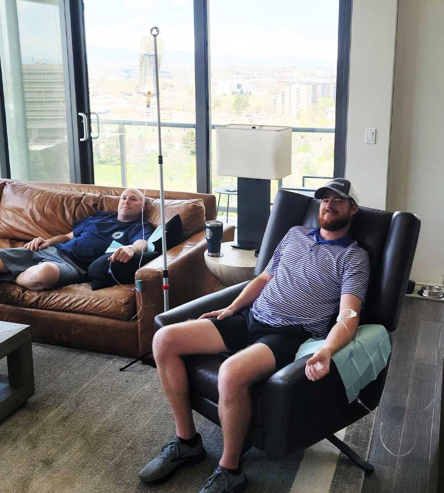 Two men relaxing and receiving mobile IV drip therapy for hangover recovery or general wellness in a luxury apartment living room with a city view.