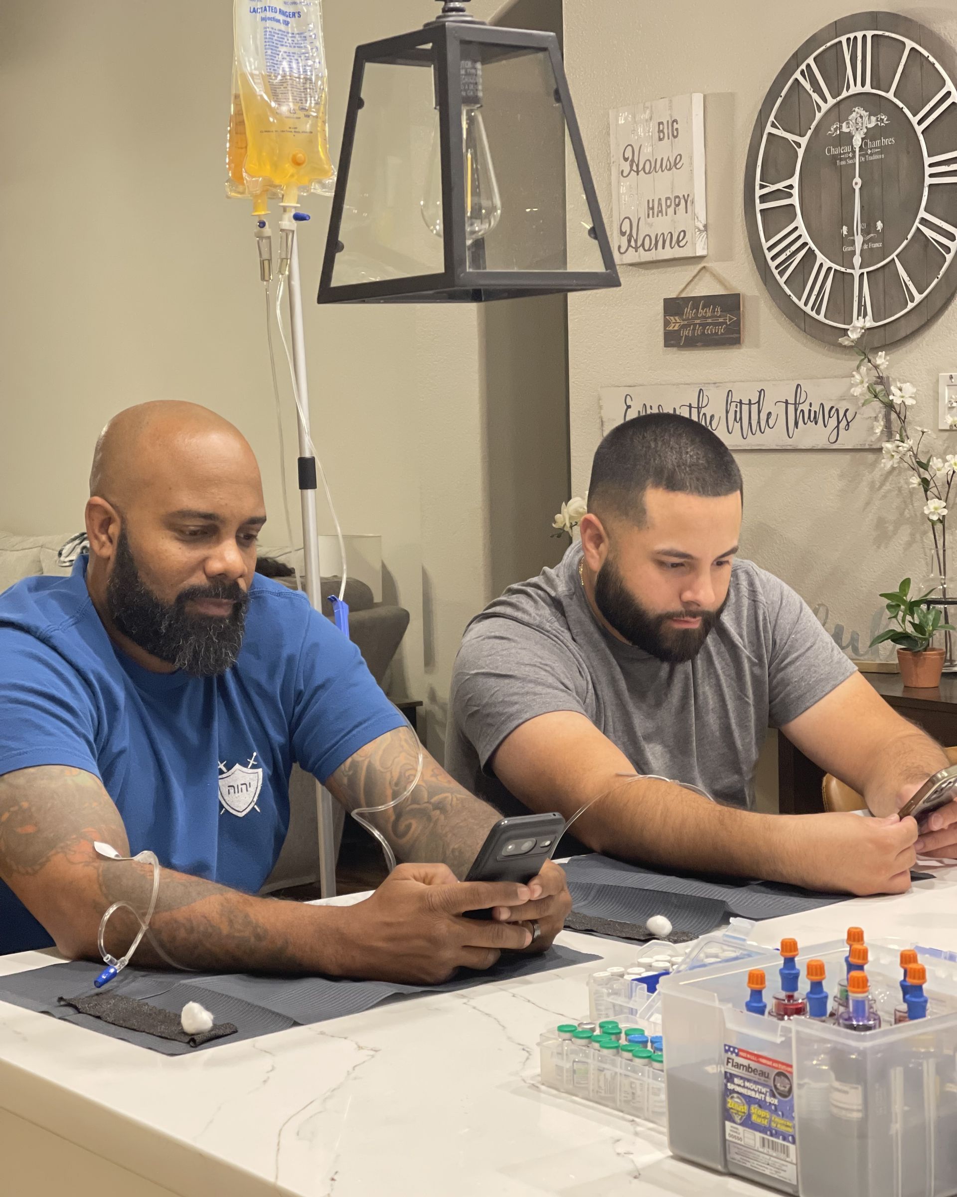 Two men sit at a counter looking at their phones, with one connected to an IV drip bag on a stand.