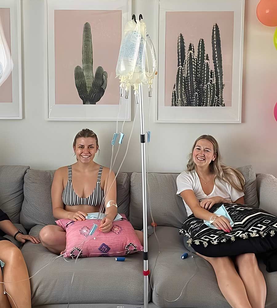 Two young women smiling and relaxing on a couch while receiving IV hydration drips during a group session.