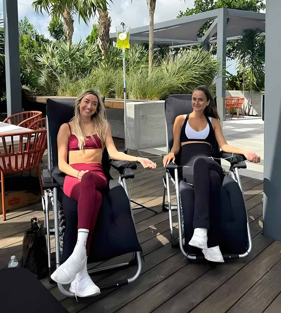 Two smiling young women in workout clothes receiving IV hydration therapy outdoors in lounge chairs on a sunny deck.