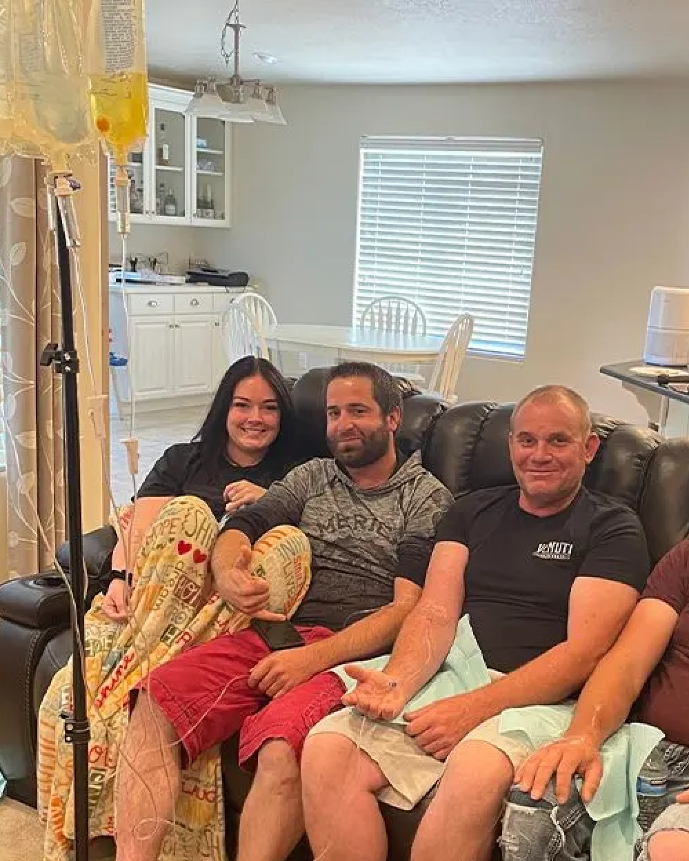 Three people sit on a black couch in a home living area, with an IV drip bag and stand positioned in the foreground.