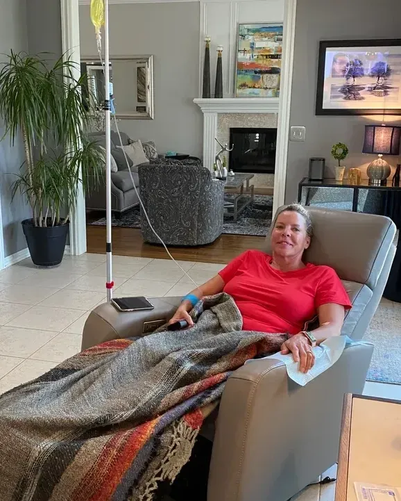 A person in a red shirt reclines in a chair at home, receiving IV therapy from a stand beside them.