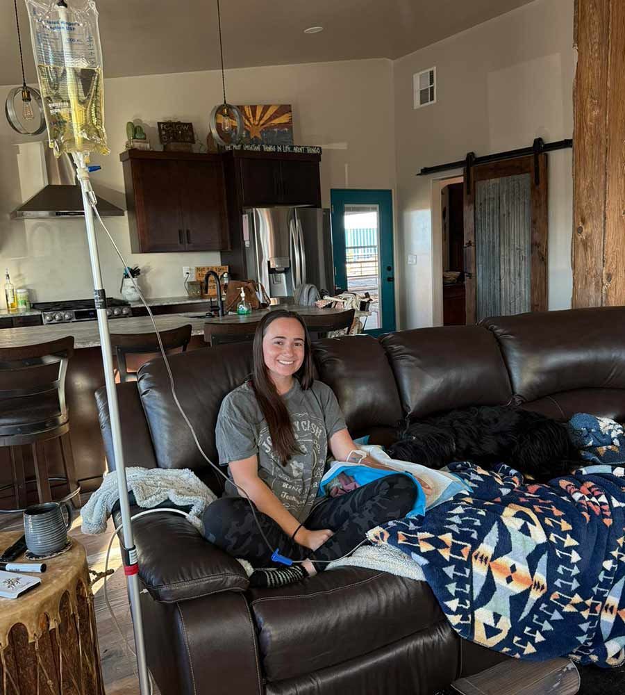 Smiling woman relaxing on a leather couch with her dog under a blanket, receiving convenient mobile IV hydration therapy in a modern, sunlit living room.