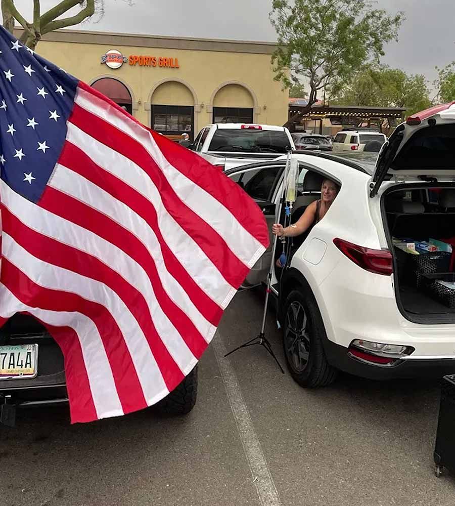Patient smiling while getting an IV infusion from the back of a car at an outdoor event or pop-up clinic.