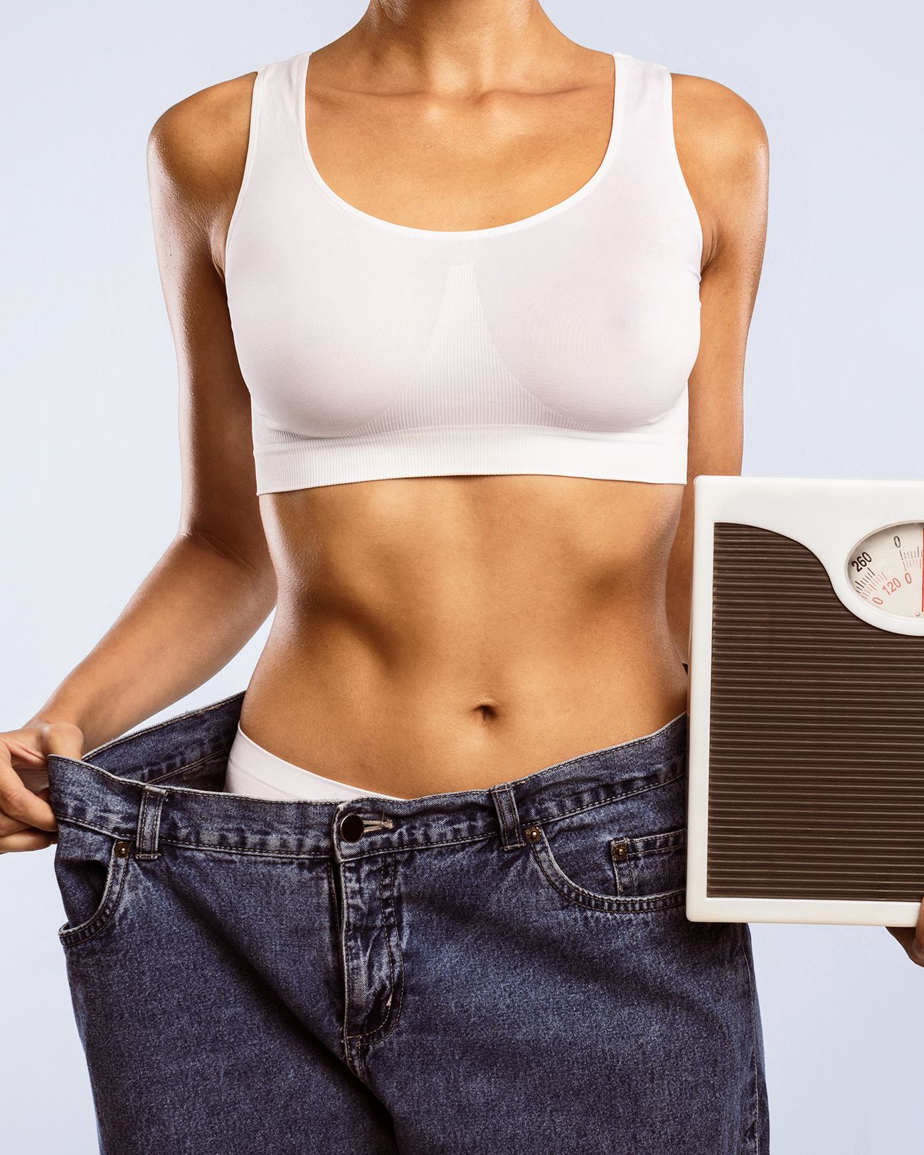 A person wearing oversized jeans holds a scale, highlighting significant weight loss against a plain background.