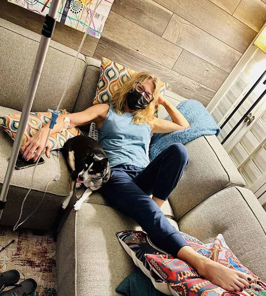 Woman wearing a face mask relaxing on a couch with her small dog while receiving an in-home IV wellness infusion.
