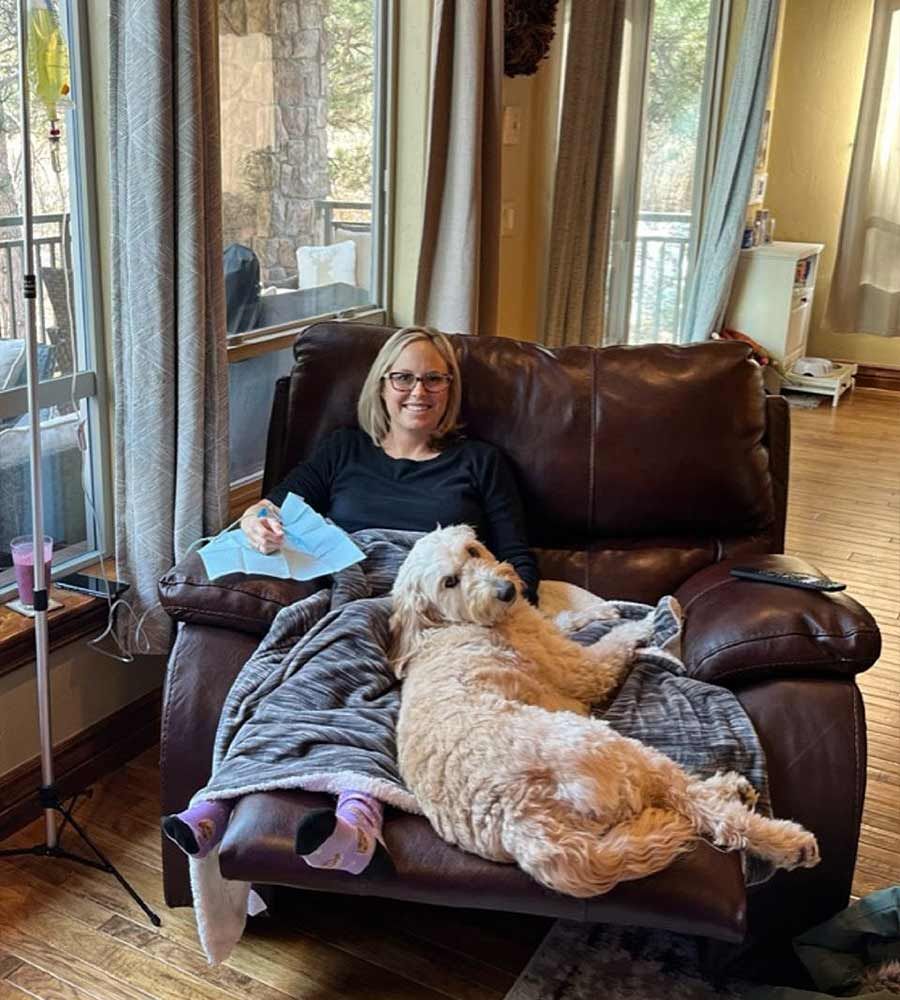 Woman in recliner with dog, receiving IV infusion. Living room setting with window.