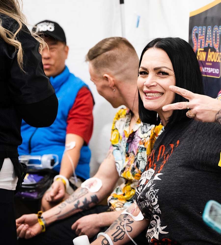 Smiling woman with arm tattoos giving a peace sign while receiving an IV hydration drip at a fun, indoor event or wellness booth.