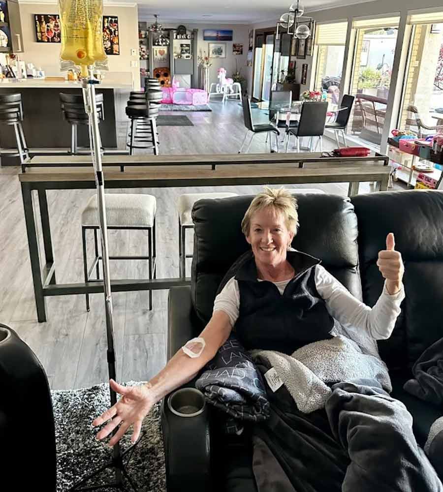 Smiling woman giving a thumbs-up while relaxing on a leather recliner and receiving mobile IV therapy at home.