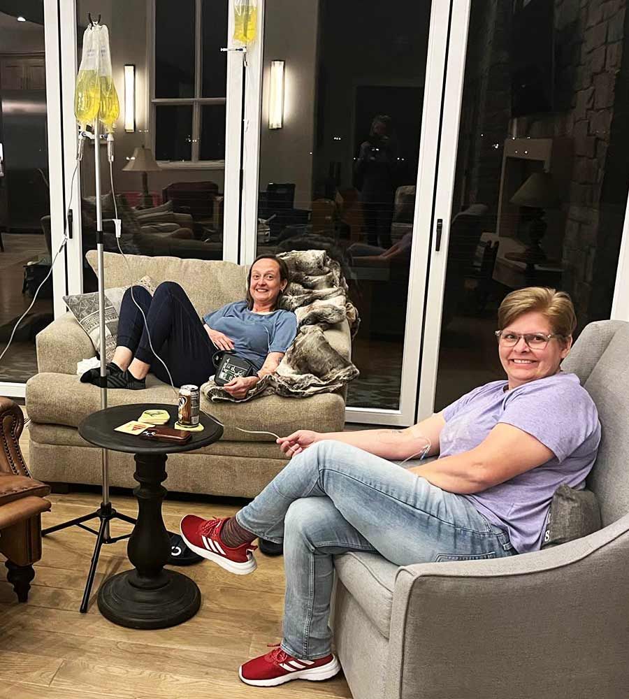 Two middle-aged women relaxing and smiling while receiving mobile IV therapy drips during an evening group session in a comfortable, modern living room.