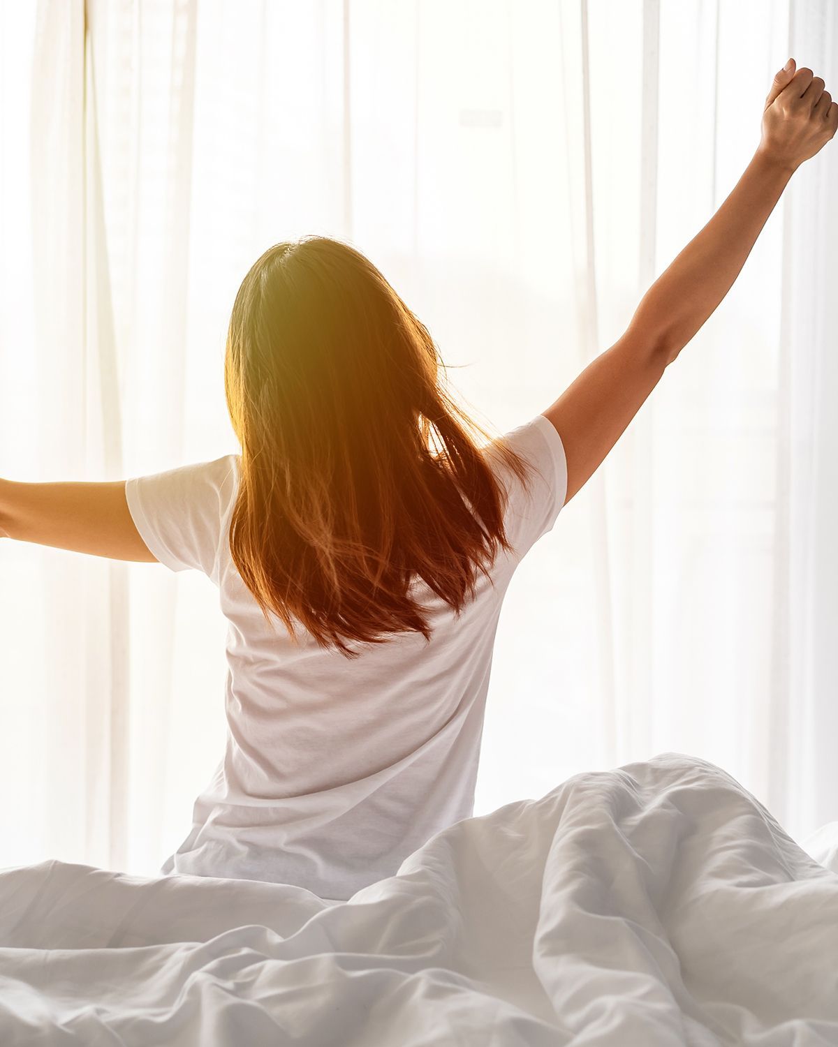 A person in a white shirt sits on a bed, back to the camera, stretching their arms toward a bright, sunlit window.