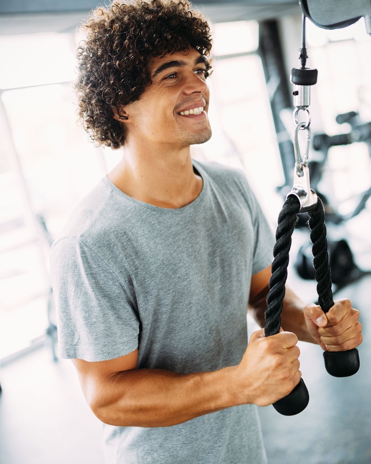 A person with curly hair smiling while performing a tricep rope pulldown exercise in a gym.