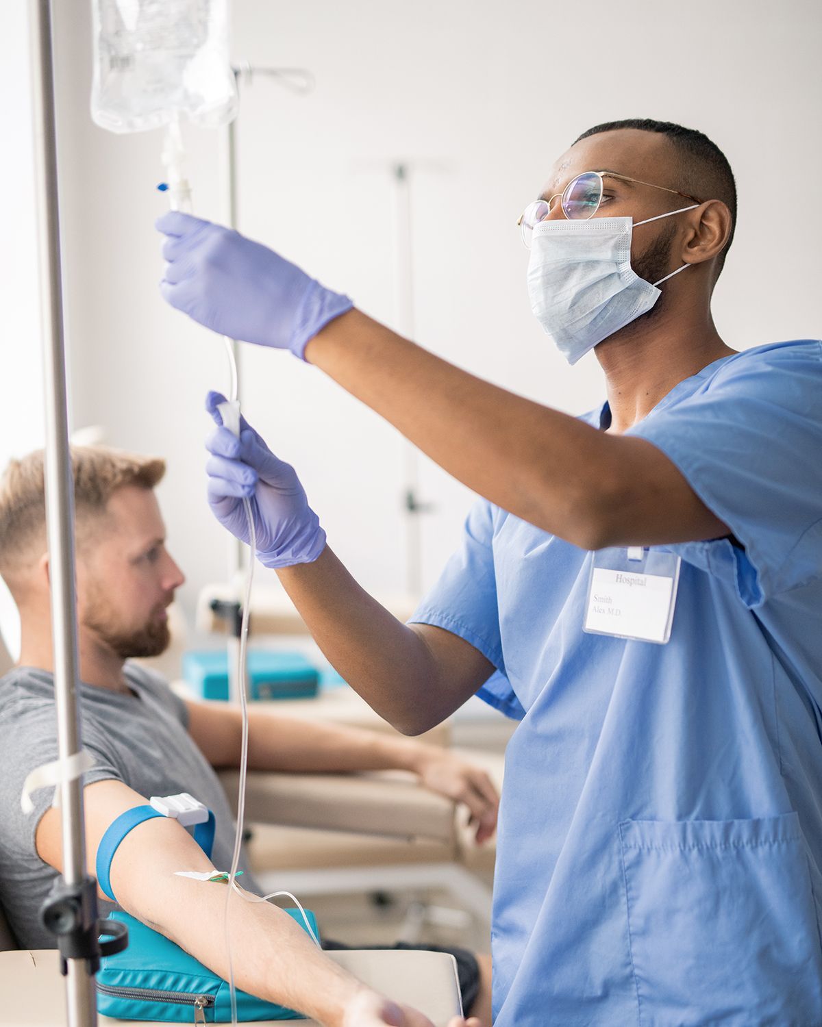 A medical professional in scrubs and a mask adjusts an IV drip for a patient in a clinical setting.