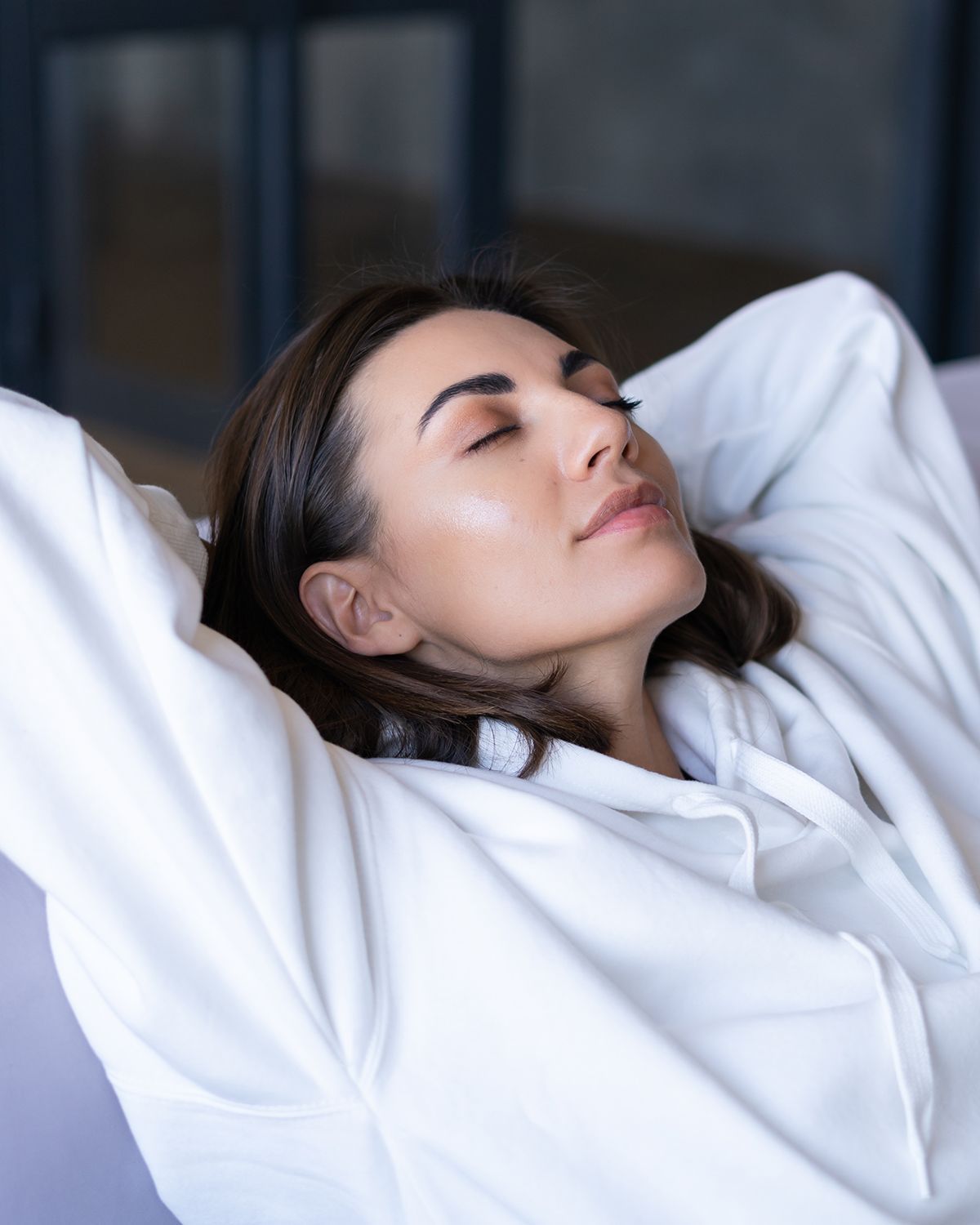 A person with dark hair wearing a white hoodie, relaxing with hands behind their head and eyes closed.