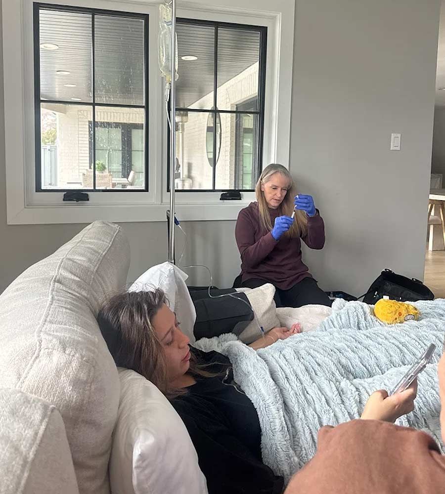 Healthcare professional (nurse wearing gloves) preparing medication while a patient receives an IV infusion in a modern living room.