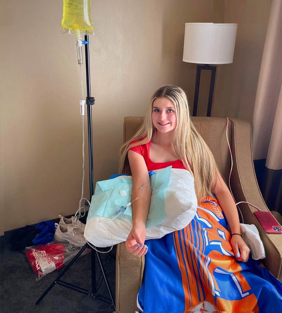 A young woman smiles while receiving an IV hydration drip infusion on a chair in a hotel room for quick recovery or wellness.