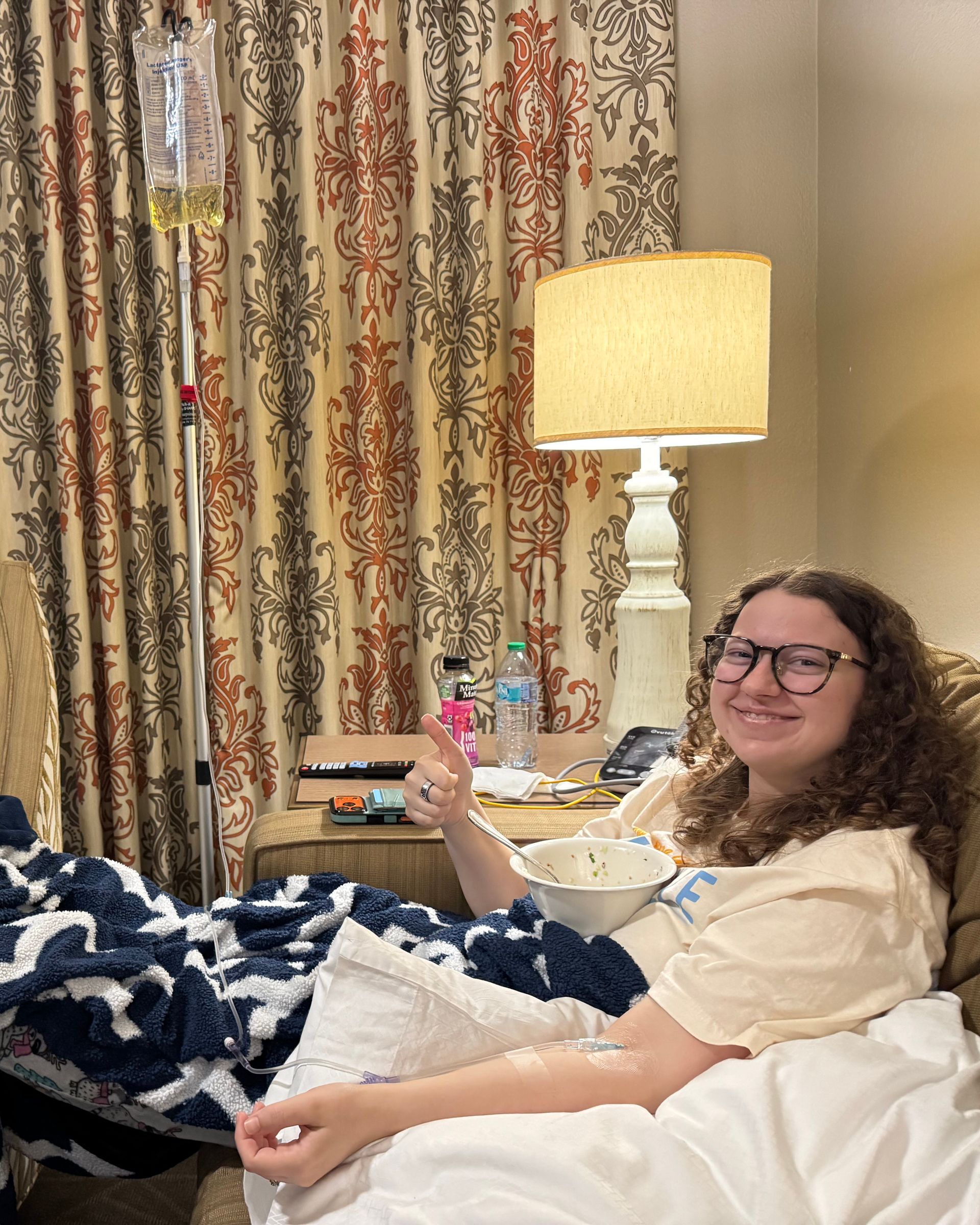 A person with curly hair and glasses smiles while giving a thumbs up, receiving IV fluids in a room with a lamp.