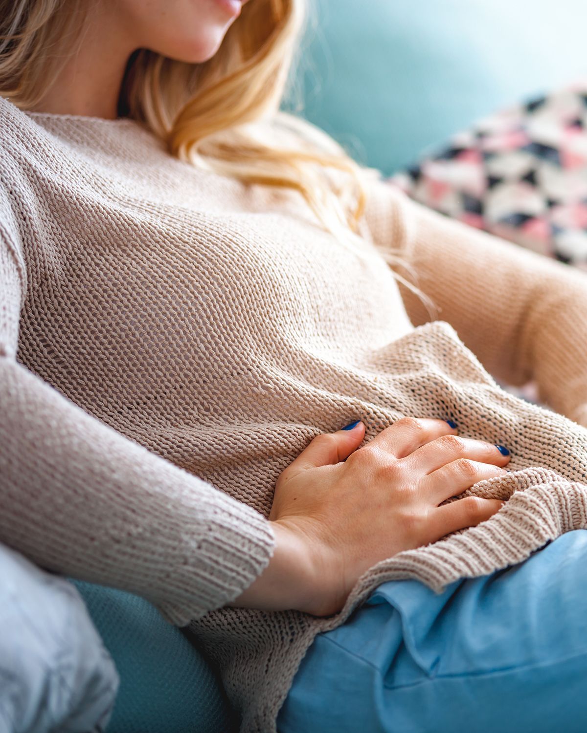 A person wearing a beige sweater resting their hand on their stomach while sitting on a couch.