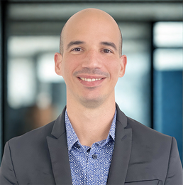 Bald man in suit jacket smiling, wearing a blue patterned shirt in an office setting.